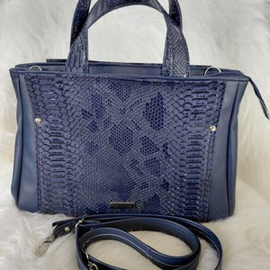 May include: A navy blue handbag with a snakeskin pattern on the front panel and handles. The bag has a matching shoulder strap with silver-tone hardware. The bag is rectangular with a zipper closure and two top handles.