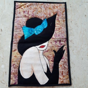 Peut inclure: Une tapisserie murale matelassée représentant une femme portant un chapeau noir avec un nœud bleu. La femme porte une robe noire et a les lèvres rouges. L'arrière-plan est un tissu beige avec un motif brun et violet.