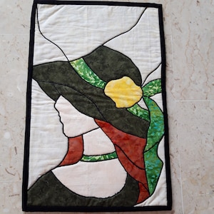 以下が含まれることがあります： 黄色の花がついた大きな緑色の帽子をかぶった女性のシルエットが描かれた、キルティングされた壁掛け。背景はクリーム色で、黒い縁取りがされている。