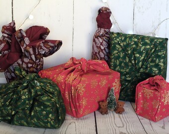 Könnte beinhalten: Mehrere Geschenke, die mit Stoff nach der Furoshiki-Methode verpackt sind. Die Stoffe weisen Muster aus goldenen Schneeflocken, Blättern und anderen Designs auf roten, grünen und bordeauxfarbenen Hintergründen auf. Ein kleiner Weihnachtsbaum und zwei Teddybären sind ebenfalls vorhanden.