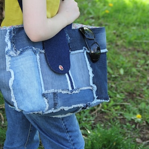 Peut inclure: Un sac cabas en jean bleu avec un design patchwork. Le sac a un bord effiloché et une bandoulière en jean bleu foncé. Des lunettes de soleil sont glissées dans le haut du sac.