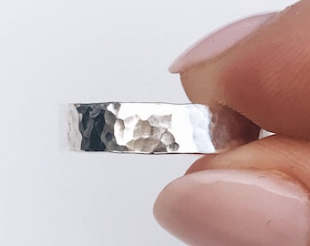 May include: A silver ring with a hammered, textured surface. The band has a flat profile and a metallic, reflective finish. The metal shows irregular indentations across the outer circumference, creating a distinct, light-catching pattern.