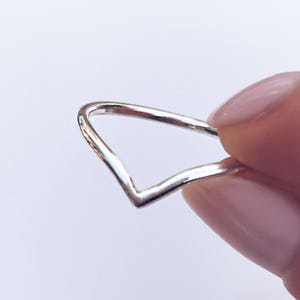 May include: A polished silver ring featuring a distinct V-shaped chevron design. The band is thin and smooth, showcasing a metallic finish that reflects light along its angular silhouette. The ring is held to display its pointed shape and structure.