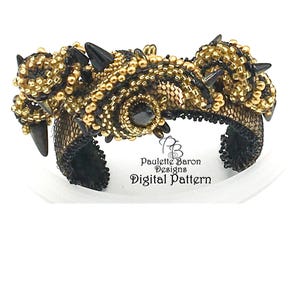 Puede incluir: Un brazalete de cuentas negras y doradas con un diseño geométrico y púas negras. El brazalete está hecho con cuentas de semillas y presenta una piedra central negra.