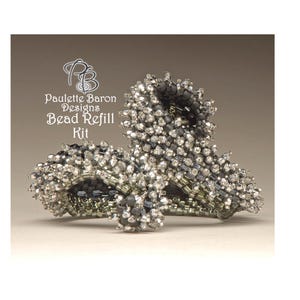 May include: A close-up of a silver and black beaded jewellery kit. The kit is made up of small, faceted beads in a variety of shades of grey and black. The beads are arranged in a complex pattern, creating a unique and eye-catching design. The kit is labelled "Paulette Baron Designs Bead Refill Kit".