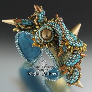 Puede incluir: Un brazalete de cuentas turquesa y dorado con una gran cuenta central de color dorado y múltiples púas. El brazalete está hecho con una variedad de cuentas, incluyendo cuentas de semillas, cuentas de bugle y cuentas redondas más grandes. Las púas son de metal y están unidas al brazalete con alambre. El brazalete es una pieza llamativa y sería perfecto para una ocasión especial.