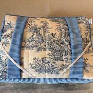 Peut inclure: Sac fourre-tout rectangulaire avec un tissu crème à motifs de scènes pastorales bleues. Il possède des poignées et des bordures en velours côtelé bleu clair. Une bande élastique blanche est tendue sur le dessus. Dimensions: 40cm x 30cm.