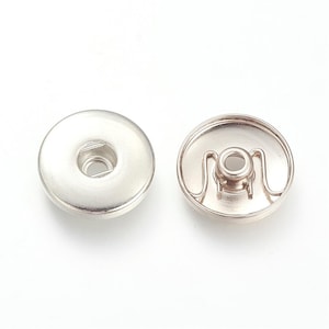 Peut inclure: Un ensemble de deux boutons-pression argentés. Le bouton de gauche est un bouton plat et rond avec un petit trou au centre. Le bouton de droite est un bouton rond avec une boucle métallique autour du trou central.