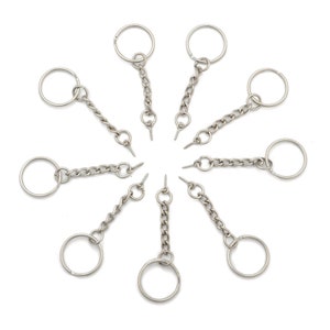 Peut inclure: Dix porte-clés argentés avec un anneau circulaire et une chaîne attachée à une vis. Les porte-clés sont disposés en cercle sur un fond blanc.