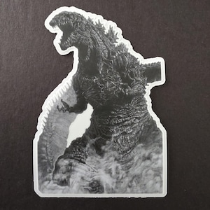 May include: A black and white sticker depicting Godzilla, a large monster, with its mouth open and smoke or mist around its feet. The sticker has a white border.