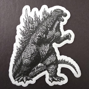May include: Black and white sticker featuring a detailed illustration of Godzilla, the iconic movie monster. The sticker shows Godzilla in a dynamic pose, with its dorsal fins and textured skin clearly visible. The sticker has a white border.
