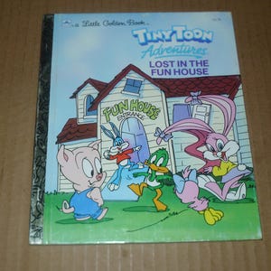 May include: A Little Golden Book titled "Tiny Toon Adventures: Lost in the Fun House." The cover features cartoon characters in front of a fun house with the words "Fun House Entrance" above the door. The book has a blue background.