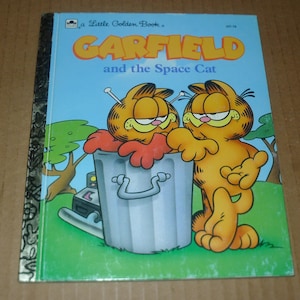 May include: A Little Golden Book titled "GARFIELD and the Space Cat" features two orange Garfield cartoon cats. One is peeking out of a silver trash can. The book has a blue cover with the title in orange and yellow.