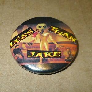 May include: A round button with a black border featuring an alien figure in a desert landscape. The words "LESS" and "THAN" are in yellow text, and "JAKE" is in yellow text below the alien.