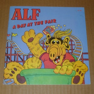 May include: A vintage ALF comic book cover titled "A Day at the Fair." The cover features a cartoon illustration of ALF on a roller coaster, holding popcorn. The background is blue with a roller coaster.
