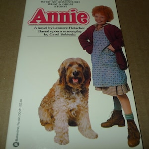 May include: The cover of the book "Annie" shows a girl with red hair and a dog. The title "Annie" is in red, with the text "LEAPIN' LIZARDS! WHAT A GIRL! WHAT AN ADVENTURE! WHAT A GREAT STORY!" above it. The book is a novel by Leonore Fleischer.