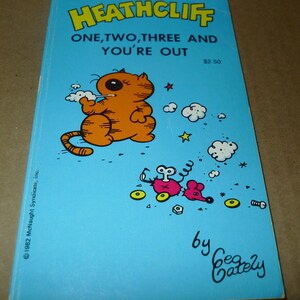 May include: A vintage paperback book titled "Heathcliff: One, Two, Three and You're Out." The cover features an orange cartoon cat smoking, with a pink mouse and other cartoon elements on a light blue background. The author's name is Gea Cately.