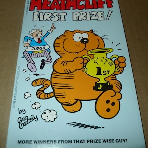 May include: A vintage comic book cover featuring Heathcliff, an orange cartoon cat, holding a trophy that says "1st". The title reads "Heathcliff First Prize!" with a cartoon judge in the background. The cover is light blue.