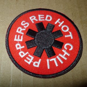 May include: Embroidered patch featuring the words "PEPPERS RED HOT CHILI" in white lettering around a black asterisk-like symbol on a red background. The patch has a black border.