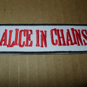 May include: Embroidered patch featuring the text "ALICE IN CHAINS" in red, stylized lettering. The text is on a white background, framed by a black border. The patch is rectangular.