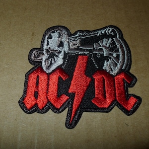 May include: Embroidered patch featuring the AC/DC logo in red, with a black border and a lightning bolt. Above the logo is a detailed illustration of a skull and cannon in gray and black. The patch is likely for clothing or accessories.