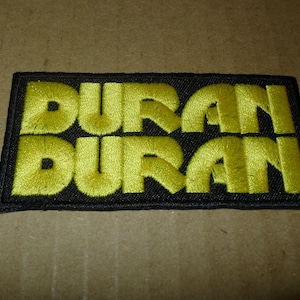 May include: Embroidered patch with the name "DURAN DURAN" in bright yellow thread on a black rectangular background. The patch has a black border and is likely for clothing or accessories.