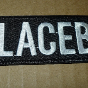 May include: Black rectangular patch with the word "PLACEBO" embroidered in white, bold, capital letters. The patch has a black border and is likely made of fabric.