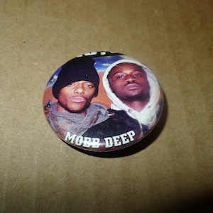 May include: Round button featuring two men, one wearing a black beanie and the other a white hooded sweatshirt. The button has the words "MOBB DEEP" in white text at the bottom. The background is a mix of orange, blue, and brown.