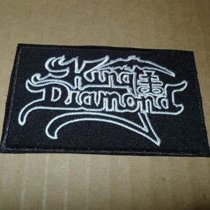 May include: Black rectangular patch with white embroidered text that reads "King Diamond". The design features stylized lettering and a mountain-like graphic above the word "Diamond".