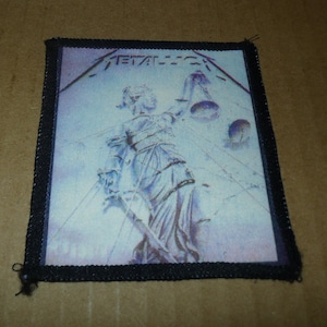 May include: A square patch featuring the Metallica logo and the Lady Justice statue. The image is in shades of blue and white, with a black border. The patch is likely for a jacket or bag.