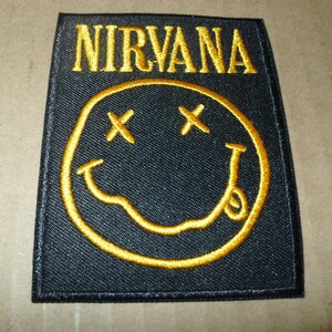 May include: Black rectangular patch with the word "NIRVANA" embroidered in gold above a yellow smiley face with crossed-out eyes and a protruding tongue. The patch is likely for clothing or accessories.