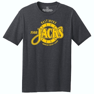 May include: A dark heather grey t-shirt with a yellow graphic of the Baltimore Jacks hockey team logo. The logo features the team name "Jacks" in a stylized font with a yellow circle around it. The text "Baltimore" is above the circle and "1988" is to the left of the circle. The text "American Hockey League" is below the circle.