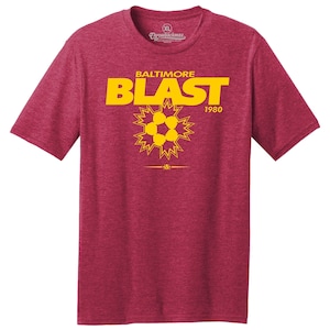 Peut inclure: Un t-shirt rouge chiné avec un motif jaune représentant un ballon de football dans un éclat d'étoile avec le texte "BALTIMORE BLAST 1980".