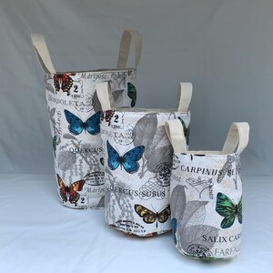 Peut inclure: Trois paniers de rangement en tissu avec des poignées crème. Les bacs cylindriques présentent un fond blanc avec un imprimé papillon et botanique. L'imprimé comprend des papillons bleus, orange et verts, ainsi que du texte.