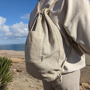 May include: A beige drawstring backpack with a front pocket and a braided drawstring. The bag is made of a textured fabric and has a small tag on the pocket. The background shows a blue sky and ocean.