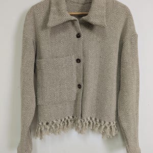 May include: A beige herringbone patterned button-up jacket with a fringe trim. The jacket has a collared neckline and long sleeves.