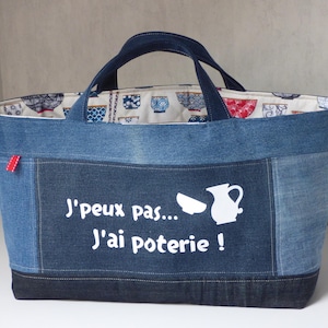 Op de afbeelding: Denim tote bag met een witte afbeelding van een kan en een kom, en de tekst "J'peux pas... J'ai poterie!". De tas heeft een patroon aan de binnenkant met blauwe en rode kommen. Voorzien van twee handvatten en een klein rood label.