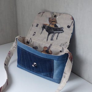 Peut inclure: Sac à bandoulière en jean avec rabat imprimé piano et notes de musique. Le sac a une forme rectangulaire avec un devant en jean bleu foncé et un intérieur de couleur claire. Une guitare est visible à l'intérieur.
