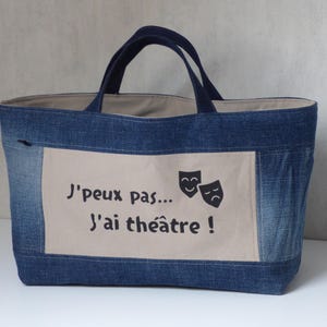 Peut inclure: Un sac fourre-tout en jean bleu avec des poignées bleu foncé. Un patch rectangulaire beige sur le devant affiche le texte "J'peux pas... J'ai théâtre !" et deux masques de théâtre. L'intérieur du sac est beige clair.