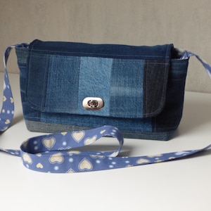 Peut inclure: Sac bandoulière en jean avec fermeture à rabat et fermoir argenté. Le sac présente un motif patchwork de différentes nuances de denim bleu. La bandoulière est bleu clair avec un motif de cœurs blancs et de petites étoiles.