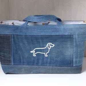 Peut inclure: Sac fourre-tout en jean avec un contour blanc de chien teckel. Le sac est de forme rectangulaire avec une poignée et un patch en jean foncé contrastant. L'intérieur est orné d'un motif de teckels.