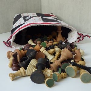 Könnte beinhalten: Eine schwarz-weiße Schachbrett-Stofftasche mit einem rot-weißen Kordelzug entleert eine Sammlung von hölzernen Schachfiguren. Die Figuren sind eine Mischung aus Schwarz, Braun und Weiß.