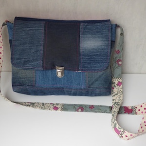Op de afbeelding: Een denim messenger bag met een patchwork design. De tas heeft een klepsluiting met een zilveren sluiting en een patroonband met roze stippen en bloemmotieven. De tas is gemaakt van verschillende tinten blauwe denim.