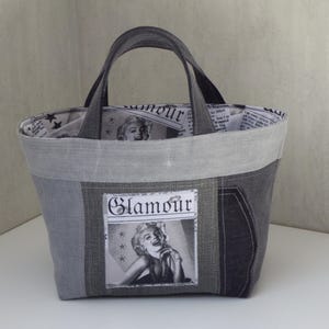 Peut inclure: Un sac fourre-tout gris avec un design patchwork. Le sac est doté d'une poignée et d'un patch rectangulaire avec le mot "Glamour" et une image en noir et blanc d'une femme. La doublure intérieure est imprimée de journaux.