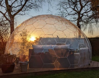 May include: A clear geodesic dome structure with a couch inside. The dome is surrounded by trees and a wooden deck. The sun is setting in the background, casting a warm glow on the scene.