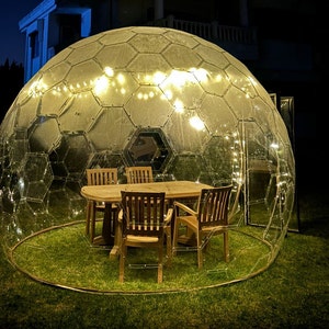 May include: A clear, geodesic dome structure illuminated by string lights, enclosing a wooden table and four chairs on a grassy area. The dome's panels create a geometric pattern, offering an outdoor dining experience. The scene is set at night.