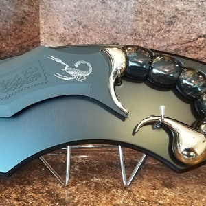 Peut inclure: Un couteau en forme de scorpion noir et argent avec une poignée noire et une lame argentée. Le couteau est monté sur une plaque en bois noir avec un support argenté.