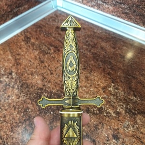 Puede incluir: Una daga dorada con grabados intrincados y un mango negro. La hoja presenta un símbolo masónico y un triángulo con un ojo.