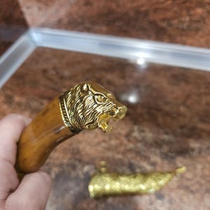 Peut inclure: Manche de couteau décoratif avec une tête de lion dorée et un manche marron clair. La tête de lion est finement détaillée, avec une crinière texturée et une gueule ouverte. La poignée est incurvée.