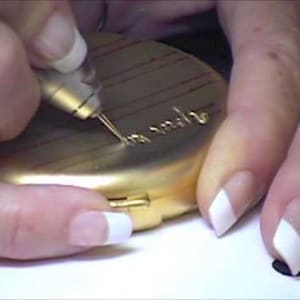 May include: A person is using a small tool to engrave a gold compact with the words "In Style".